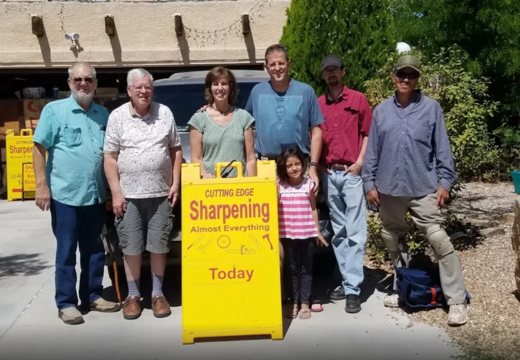 6 people standing by a sign ‘CUTTING EDGE Sharpening Almost Everything Today’, a sharpening service.