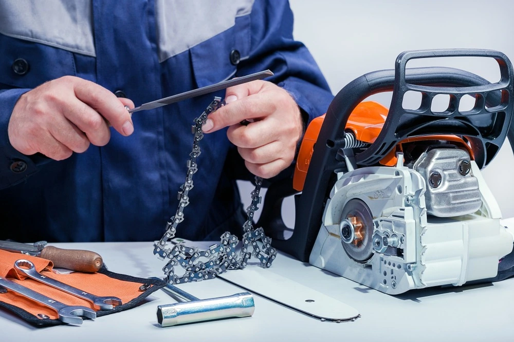 Person sharpening a chainsaw chain with a file next to the chainsaw and tools.